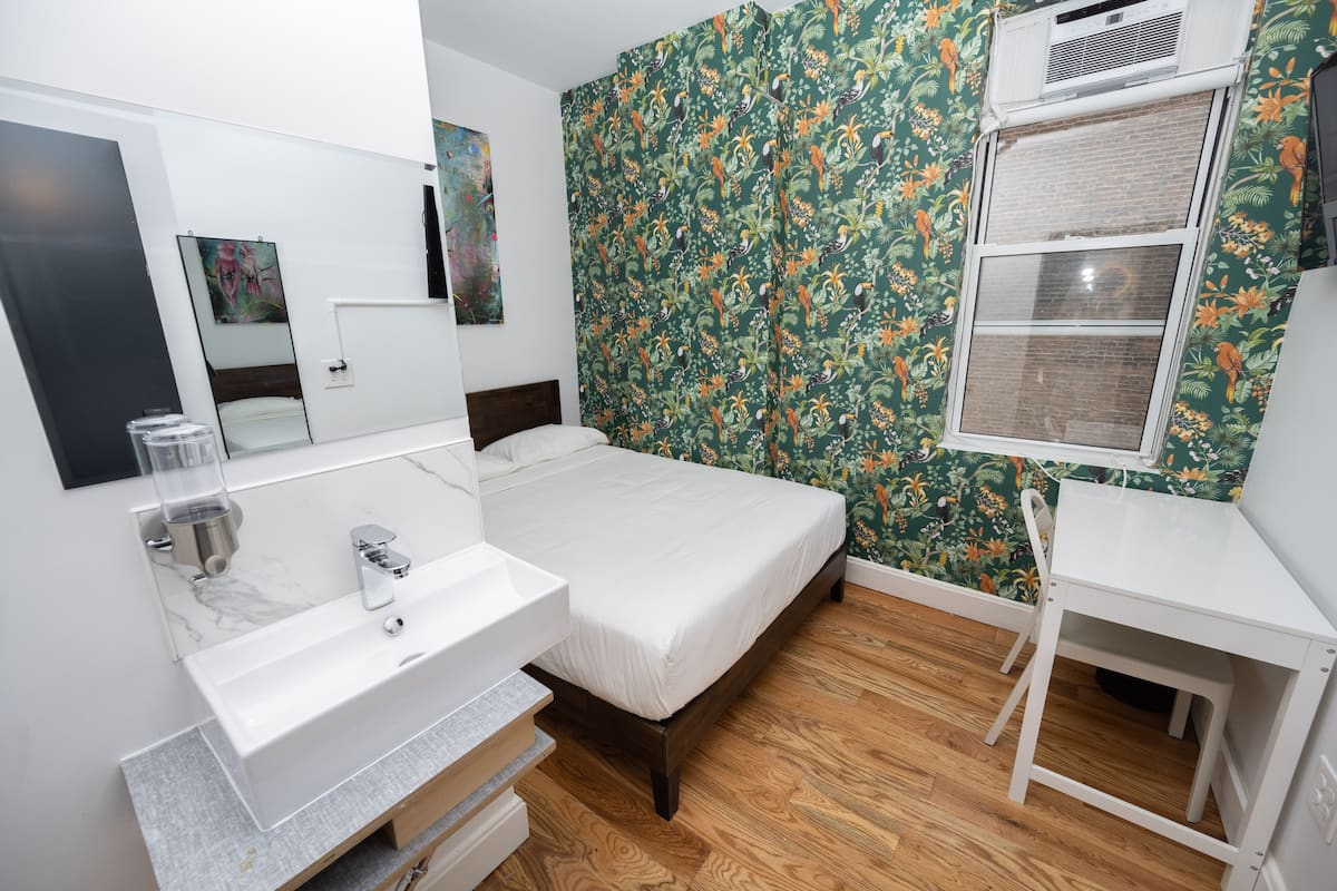 A bedroom with a bed, a white desk, a mirror, a sink, and a window with a floral wallpaper.