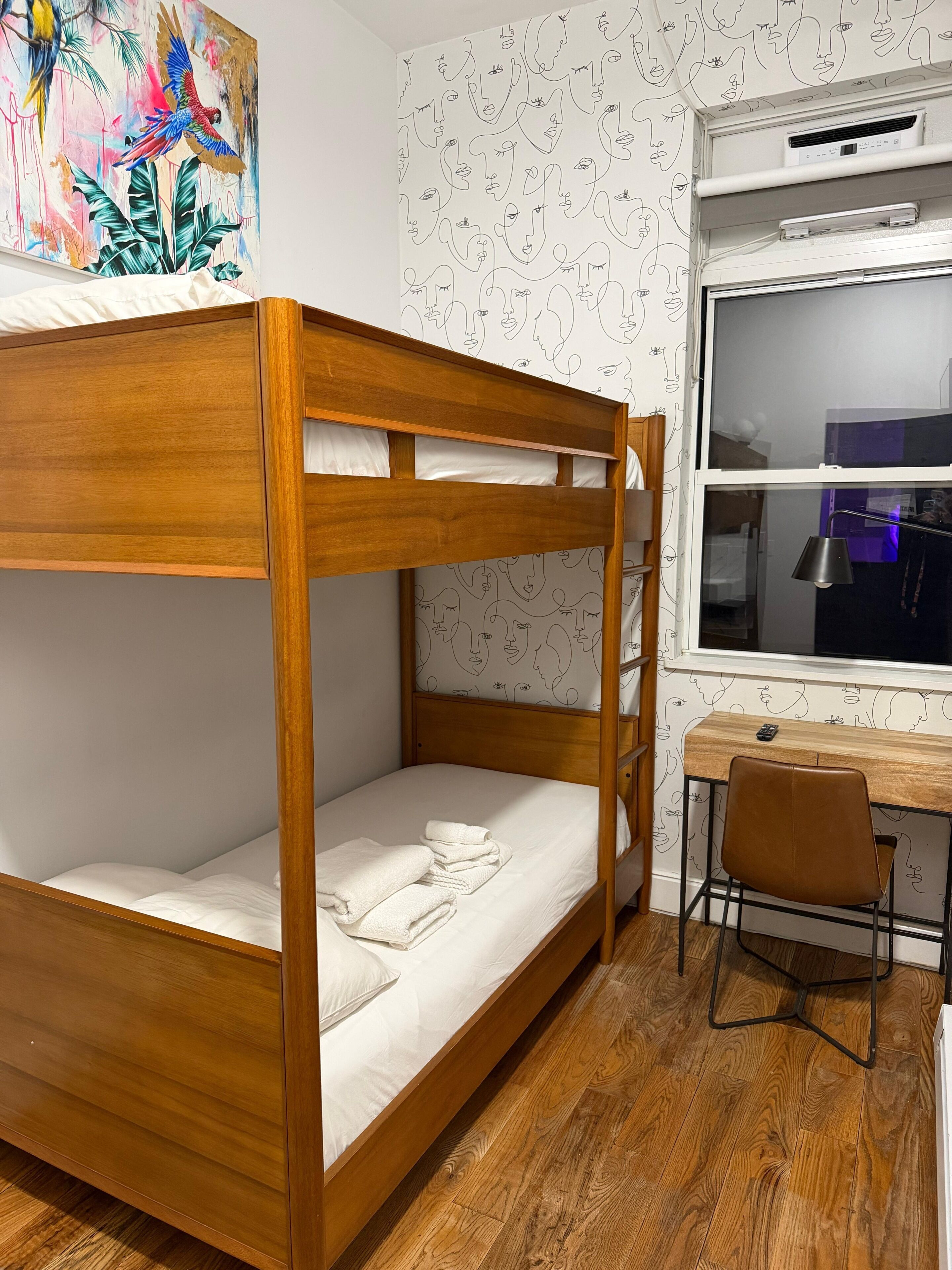 Un lit superposé avec un bureau et une chaise, une fenêtre et un mur orné d’un motif représentant un visage.