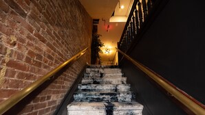 A staircase with a brick wall on one side and a dark wall on the other, leading to a room with a chandelier. - The Flat NYC (New York)