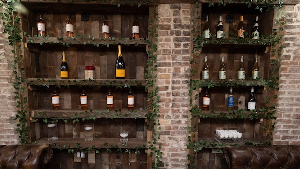 A bar area with wooden shelves displaying various bottles, a glass display case, and two leather sofas. - The Flat NYC (New York)
