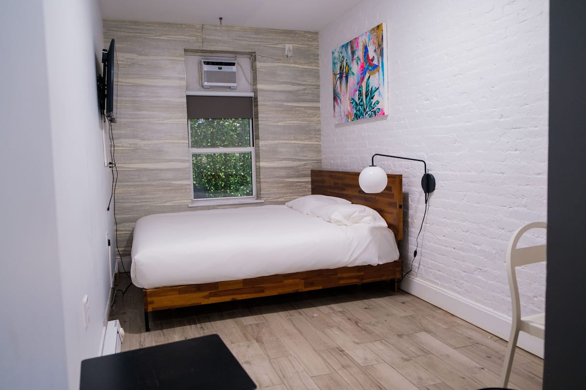 A bedroom with a wooden bed, a white wall, a window, and a colorful painting.