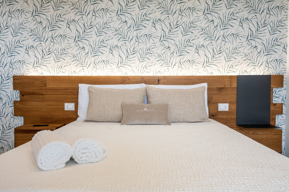 Una cama bien tendida, con cabecera de madera, dos almohadas color beige y toallas blancas enrolladas al lado.