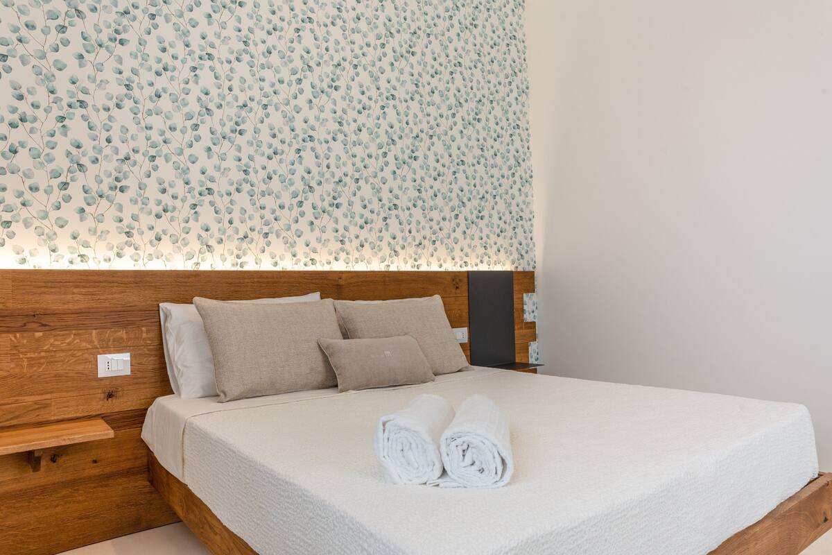 Una cama bien hecha con una colcha blanca y dos toallas enrolladas, un cabecero de madera y una pared con un estampado de hojas.