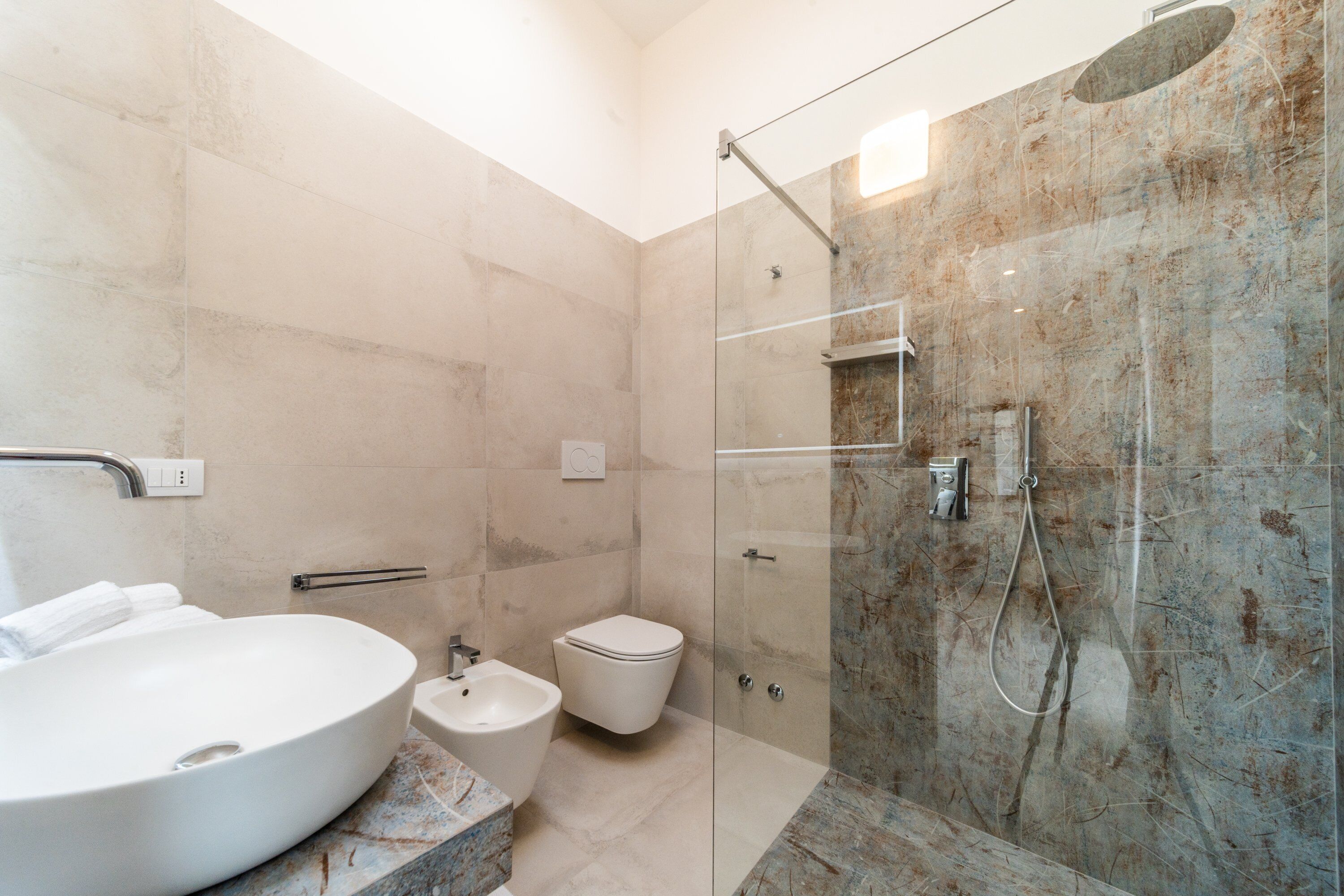 Une salle de bain moderne dotée d’un grand lavabo ovale, d’un bidet, de toilettes et d’une douche séparée par une paroi en verre.