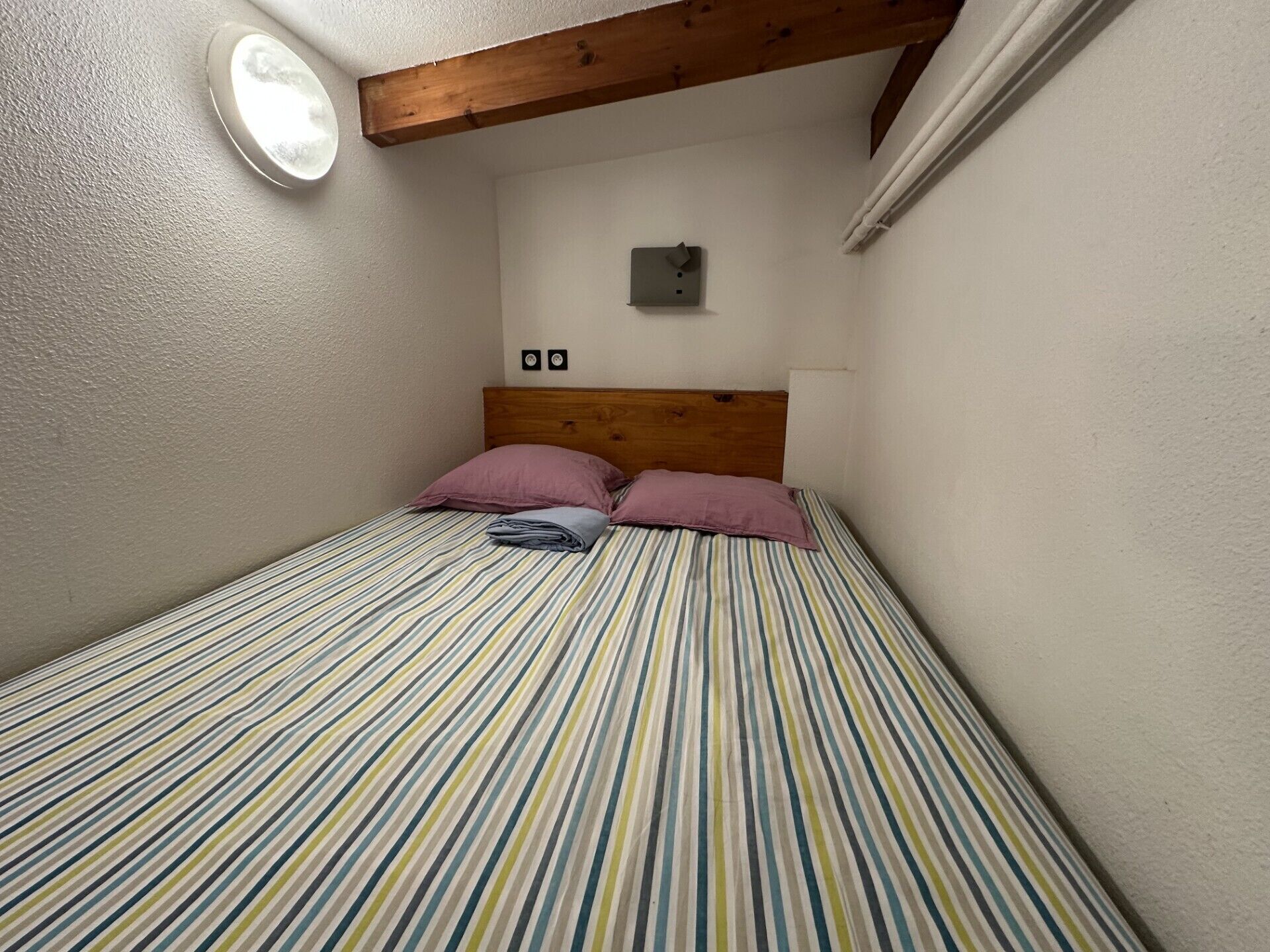A small, single-bed room with a wooden headboard, striped bedding, and a wall-mounted light fixture.