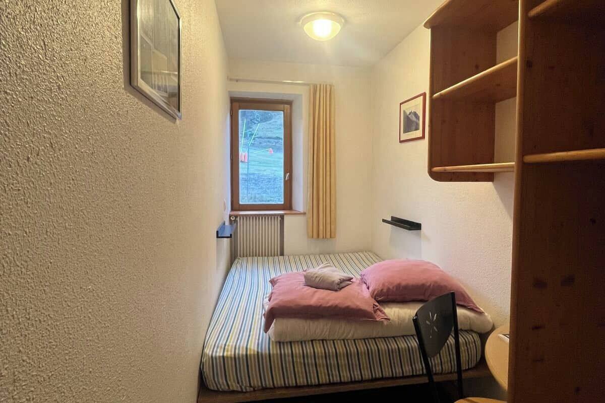 A small, narrow room with a single bed, a window with curtains, and a wooden shelf unit.