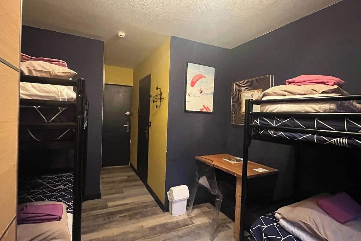 A dormitory room with bunk beds, a desk, and a door leading to another room.