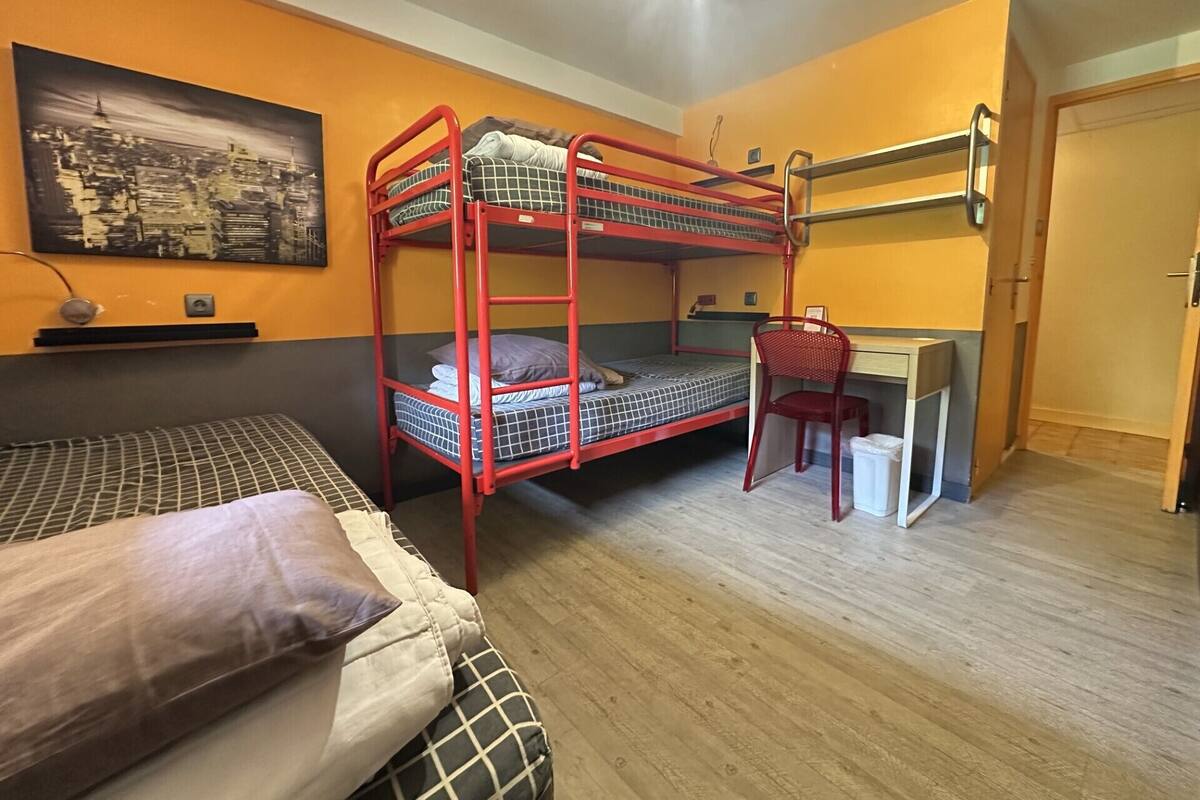 A dormitory room with bunk beds, a desk, a chair, and a bed with a patterned bedspread.