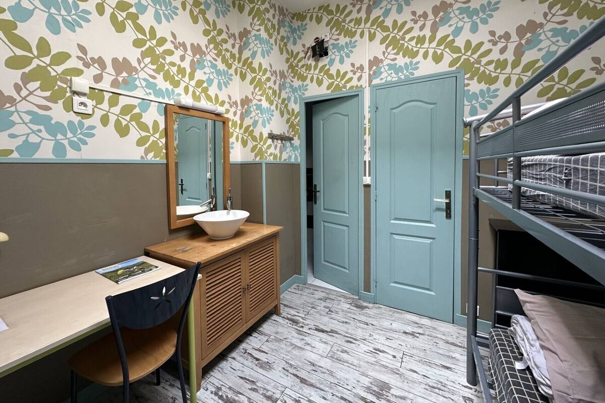 A compact bedroom with a bunk bed, a bedside table, a chair, and a bathroom with a sink and mirror.