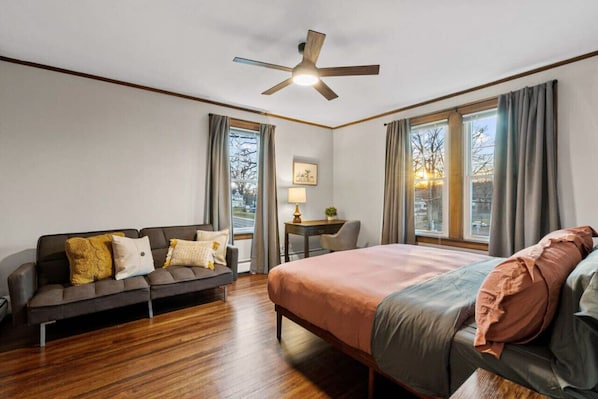 A hotel room with a bed, a sofa, a desk, a chair, and large windows.