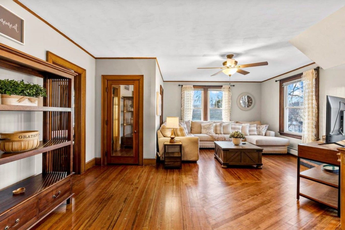 A spacious living room with a wooden floor, a ceiling fan, a sofa set, a coffee table, and a television.