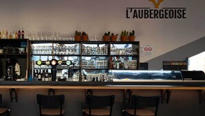 Een bar met een bord waarop 'L'Aubergeoise' staat, planken vol glazen en flessen, en drie hangende lampen.