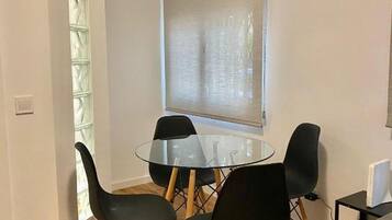 Een klein zithoekje met een glazen tafel en vier stoelen, een raam met zonwering, en een wandairconditioner.