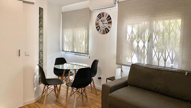 A modern living room with a sofa, a dining table with chairs, a wall clock, and a window with blinds. - Mirasierra Center - Metro Usera 02 (1 bedroom apartment) (Madrid)