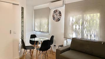 Een moderne woonkamer met een bank, een eettafel met stoelen, een wandklok en een raam met jaloezieën.