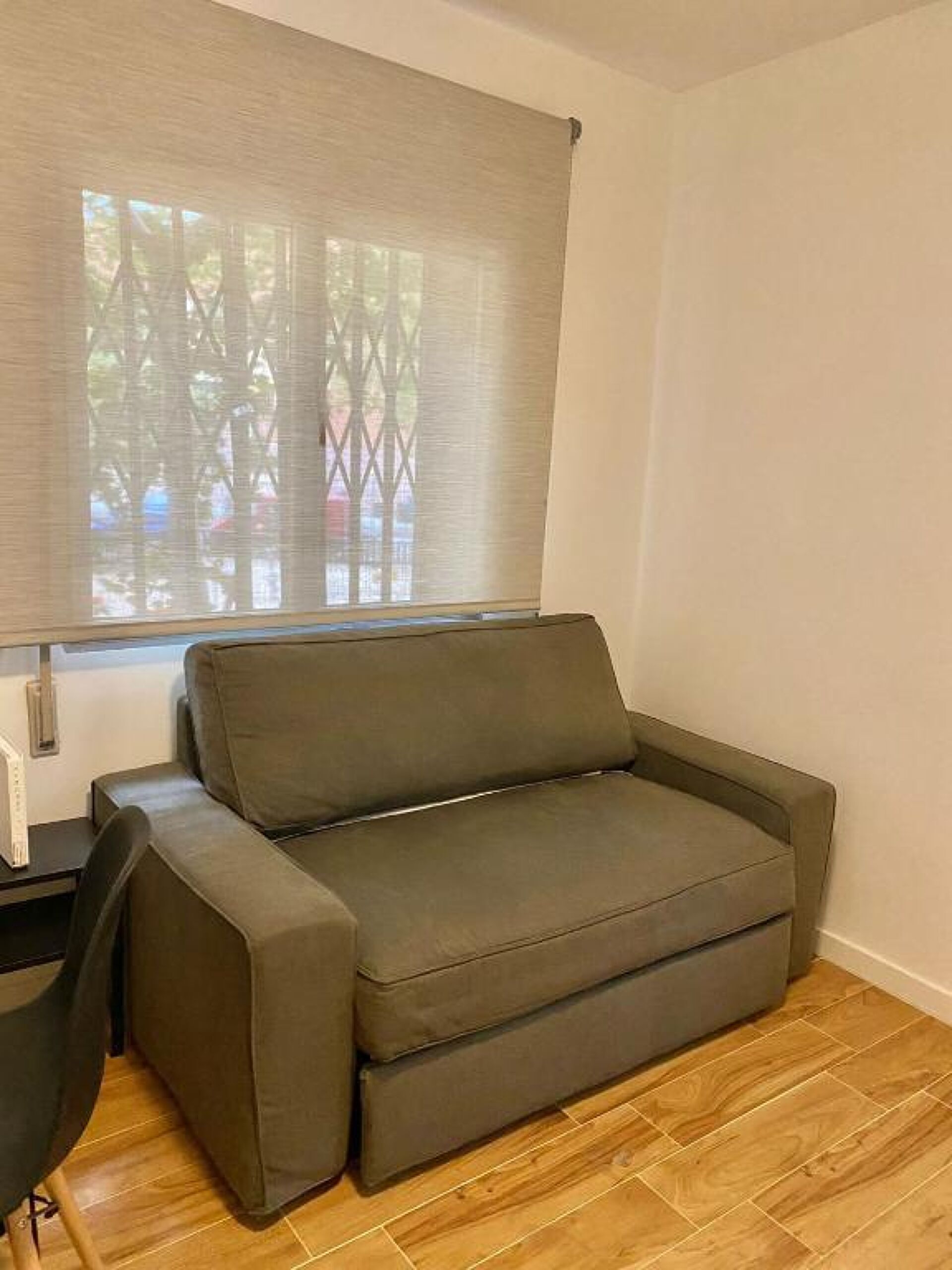 Een kamer met een grijze bank, een klein tafeltje en een raam met zonwering.