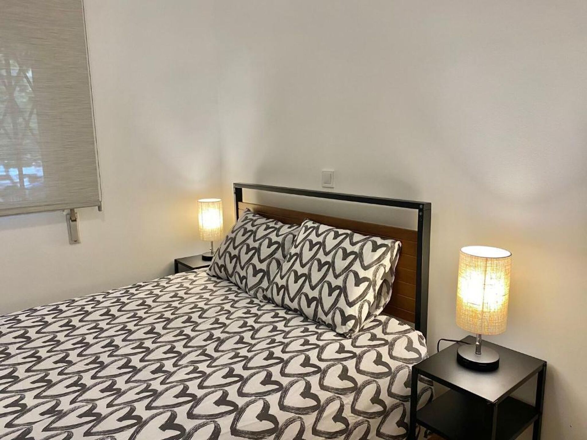 Un lit avec une couvre-lit à motifs, une tête de lit, deux lampes de chevet et une fenêtre avec des stores.