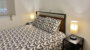 A bed with a patterned bedspread, a headboard, two bedside lamps, and a window with blinds. - Mirasierra Center - Metro Usera 02 (1 bedroom apartment) (Madrid)