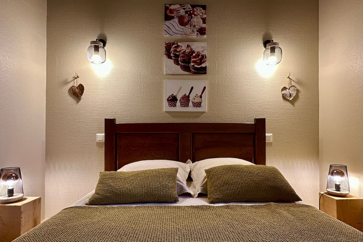A neatly made bed with a wooden headboard, two green pillows, and a textured blanket. There are wall-mounted light fixtures on either side of the bed, and two heart-shaped decorations hanging on the wall.