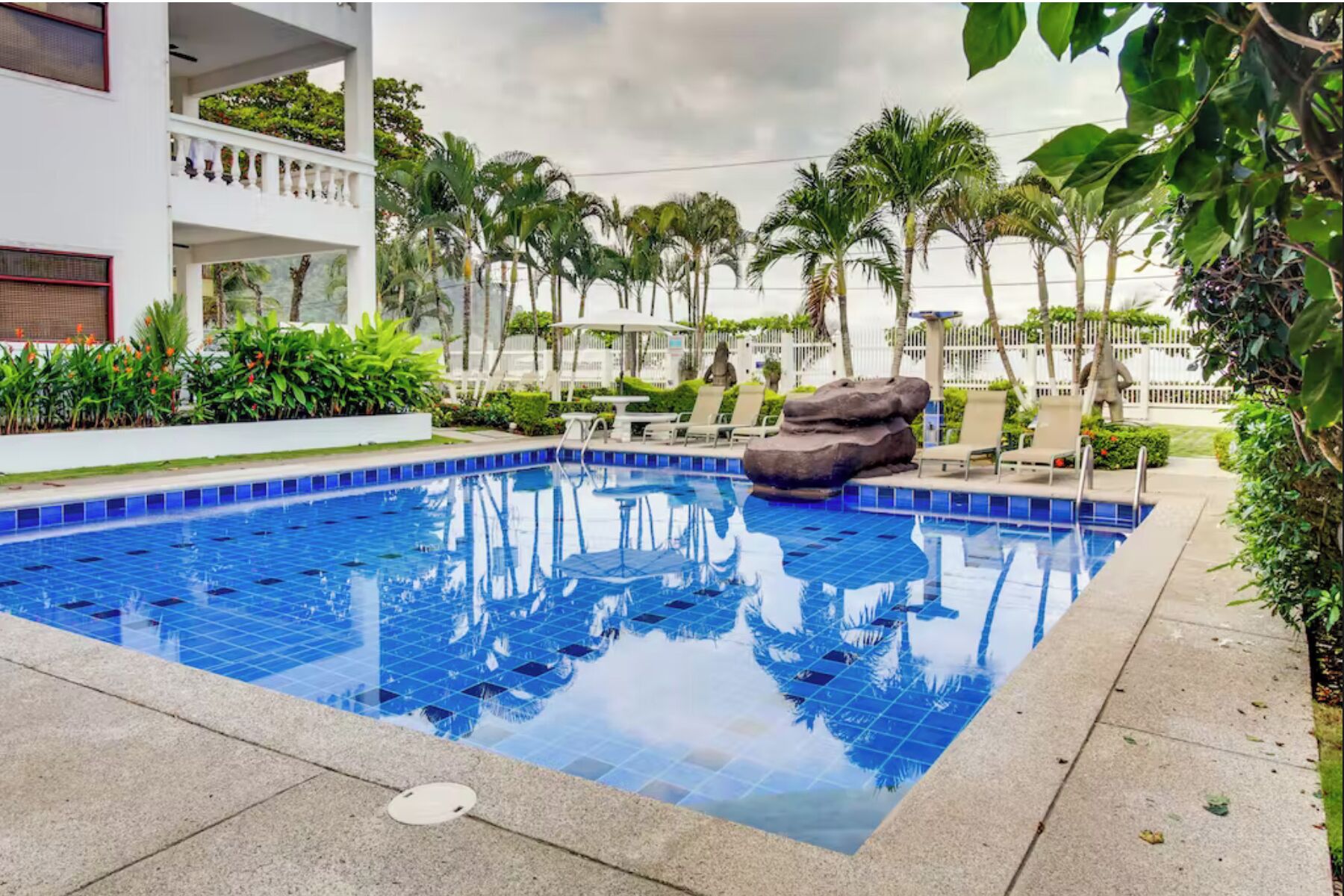 A swimming pool with blue tiles, surrounded by palm trees and lounge chairs.