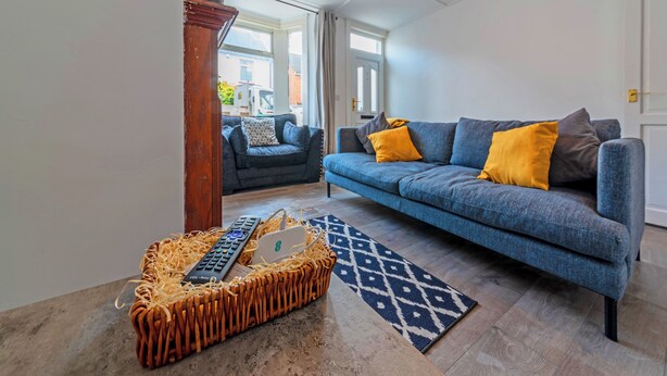 Una sala de estar con un sofá azul, un tapete estampado y una cesta de mimbre con controles remotos.