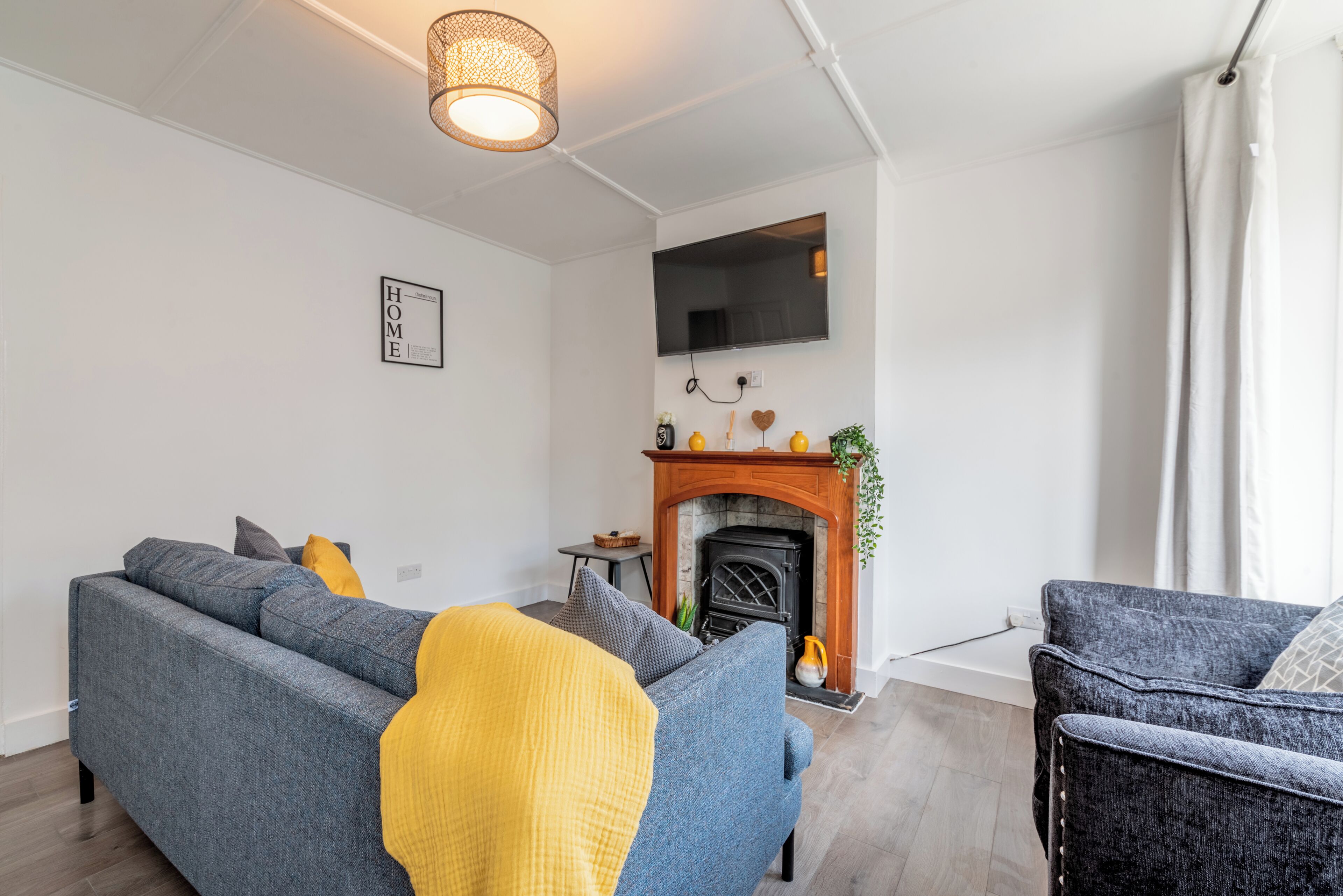 Een moderne woonkamer met een grijze bank, een gele plaid, een open haard en een flatscreen televisie aan de muur.