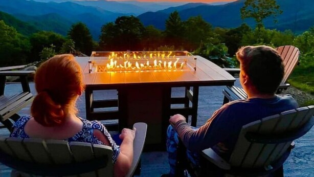 Due persone sedute a un tavolo con un sistema integrato di fuoco, con vista su un paesaggio montuoso al tramonto.