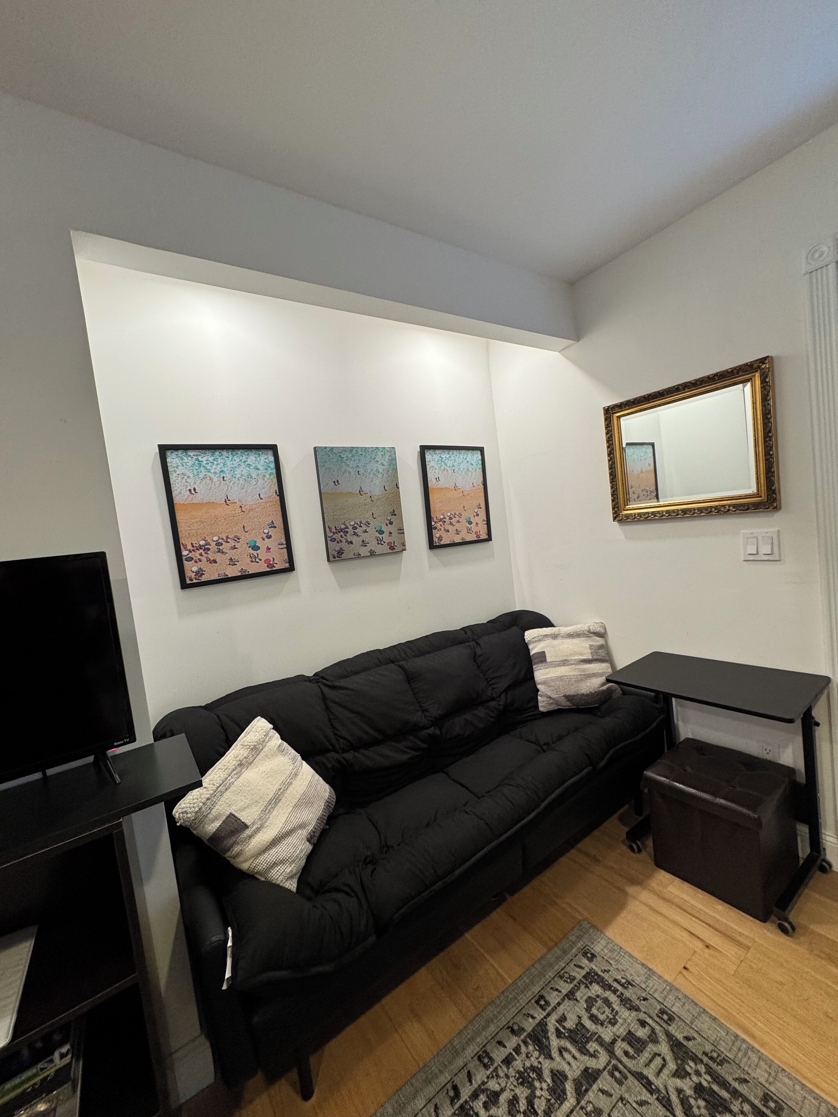 Un salon moderne avec un canapé noir, un téléviseur à écran plat et trois tableaux encadrés représentant des scènes de plage, accrochés au mur.