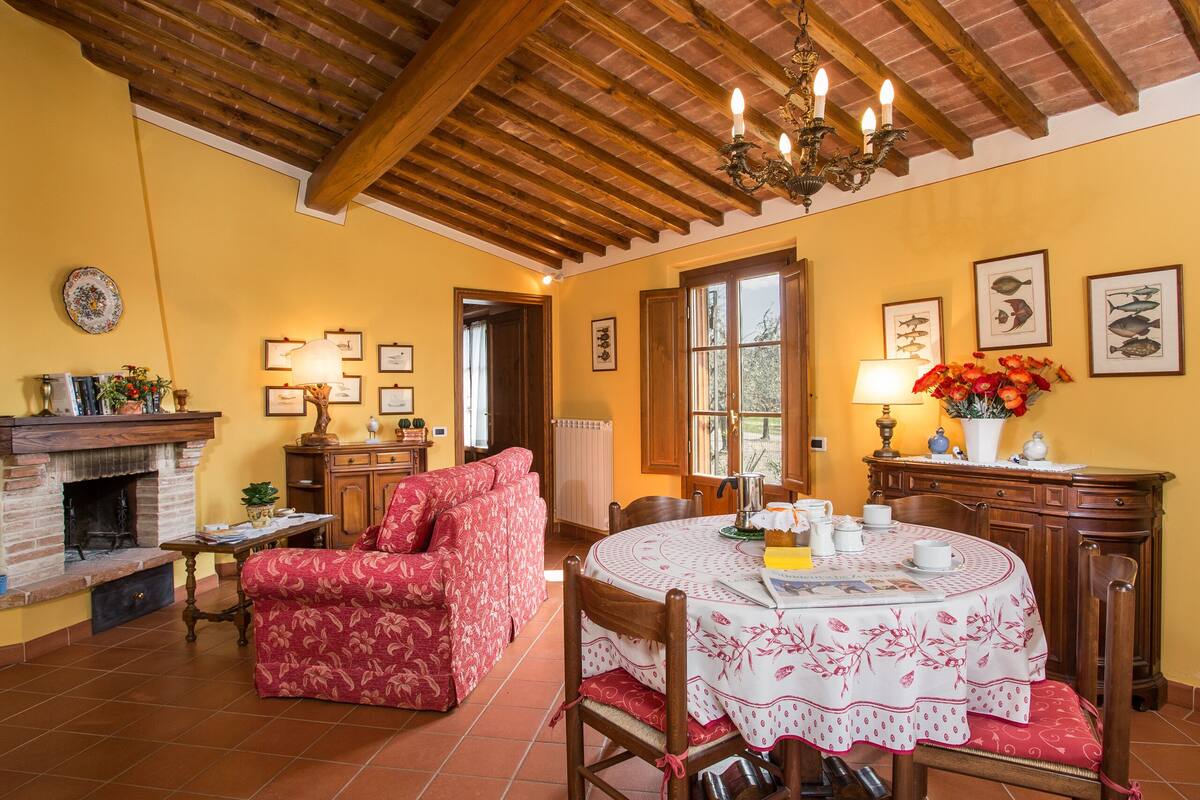Uma sala de estar acolhedora com lareira, uma mesa de jantar com toalha de mesa florida e um sofá vermelho estampado.