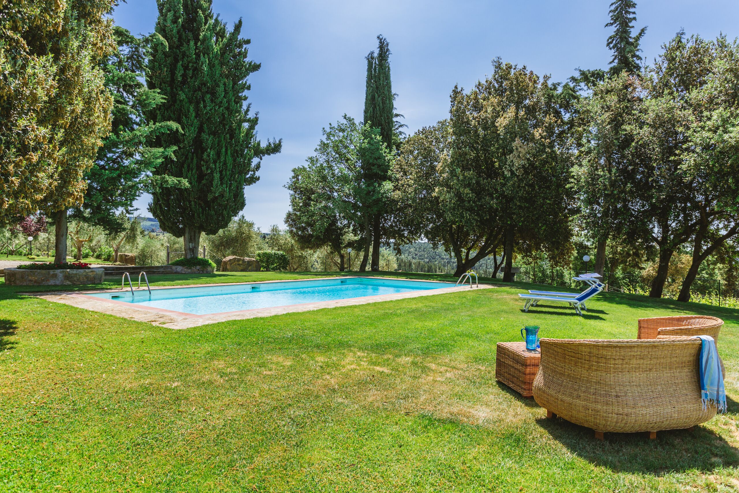 A swimming pool surrounded by a grassy lawn, with a wicker lounge chair and a sun lounger in the background.