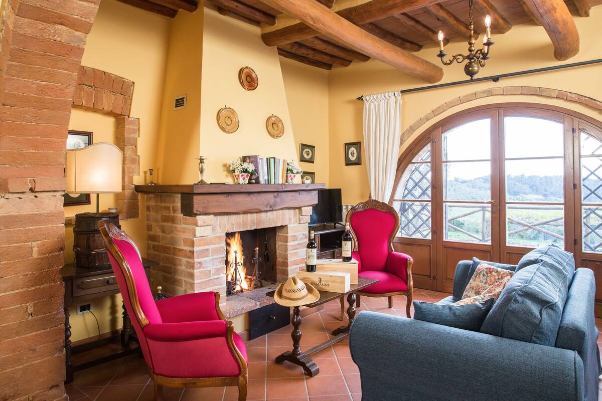 Uma sala de estar acolhedora com lareira, móveis vermelhos e azuis, e uma grande janela em arco que oferece uma vista panorâmica.