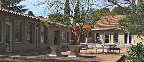 Un patio con una mesa y sillas, un edificio con techo de tejas y un árbol de hojas verdes.