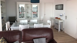 A dining area with a glass table, white chairs, a brown leather sofa, a white cabinet with glass doors, and a blue pendant light. - LES BALCONS ROUGES Face Mer - Impressive sea view (SAINTE CECILE)