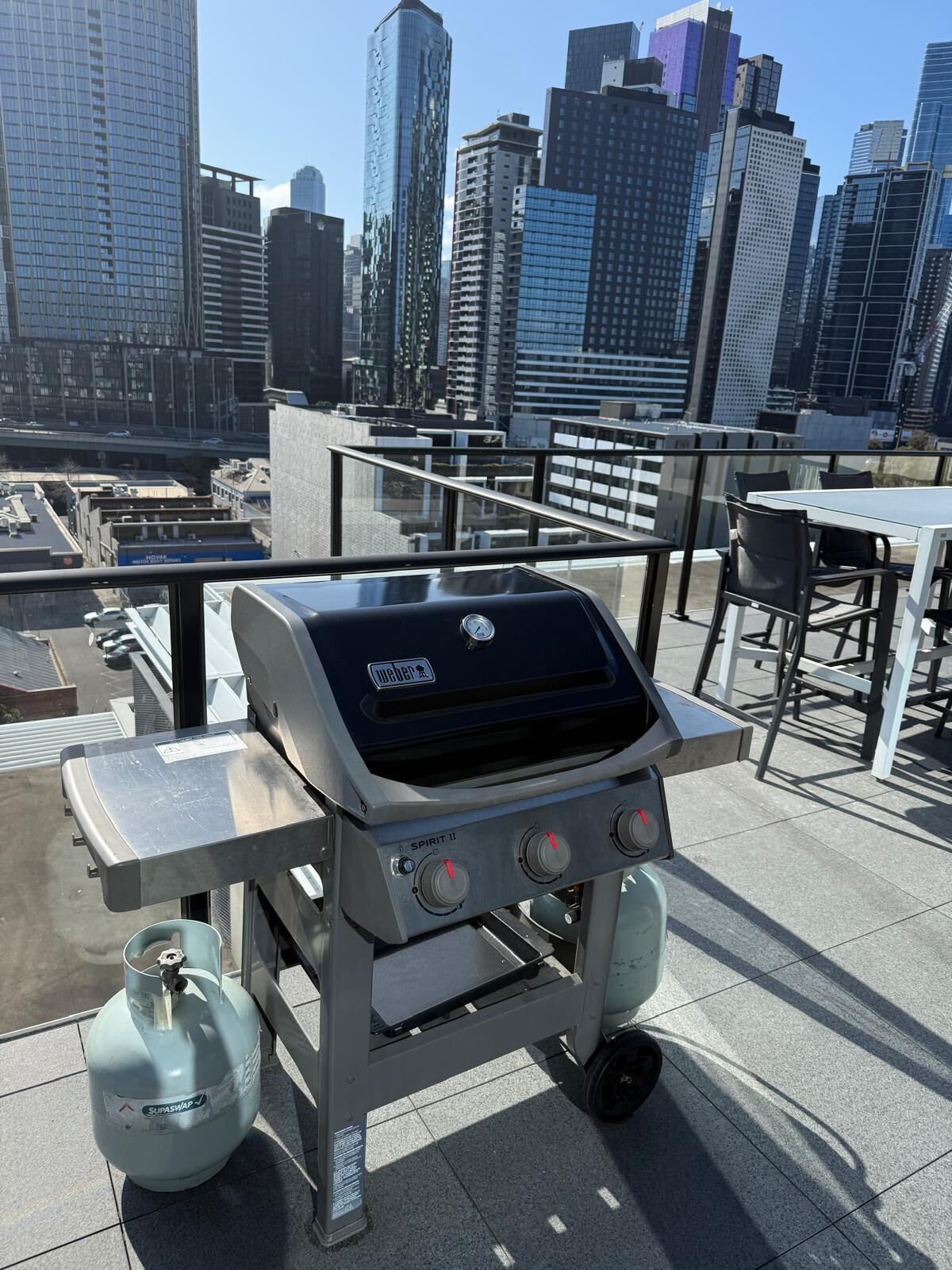 A rooftop grill with a gas tank, a table, and chairs, overlooking a cityscape.