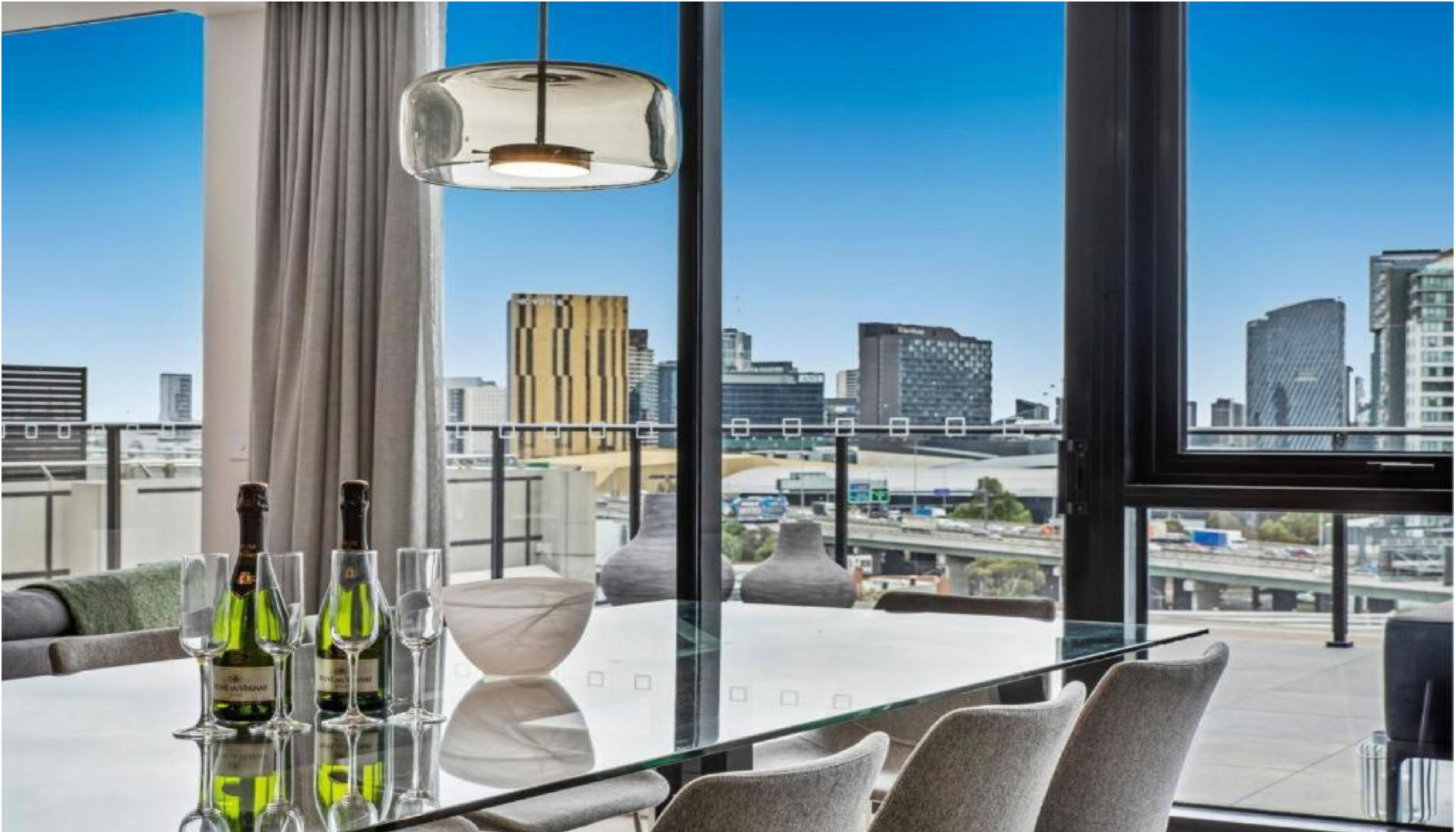 A modern dining area with a city view, a glass table, and three bottles of wine.