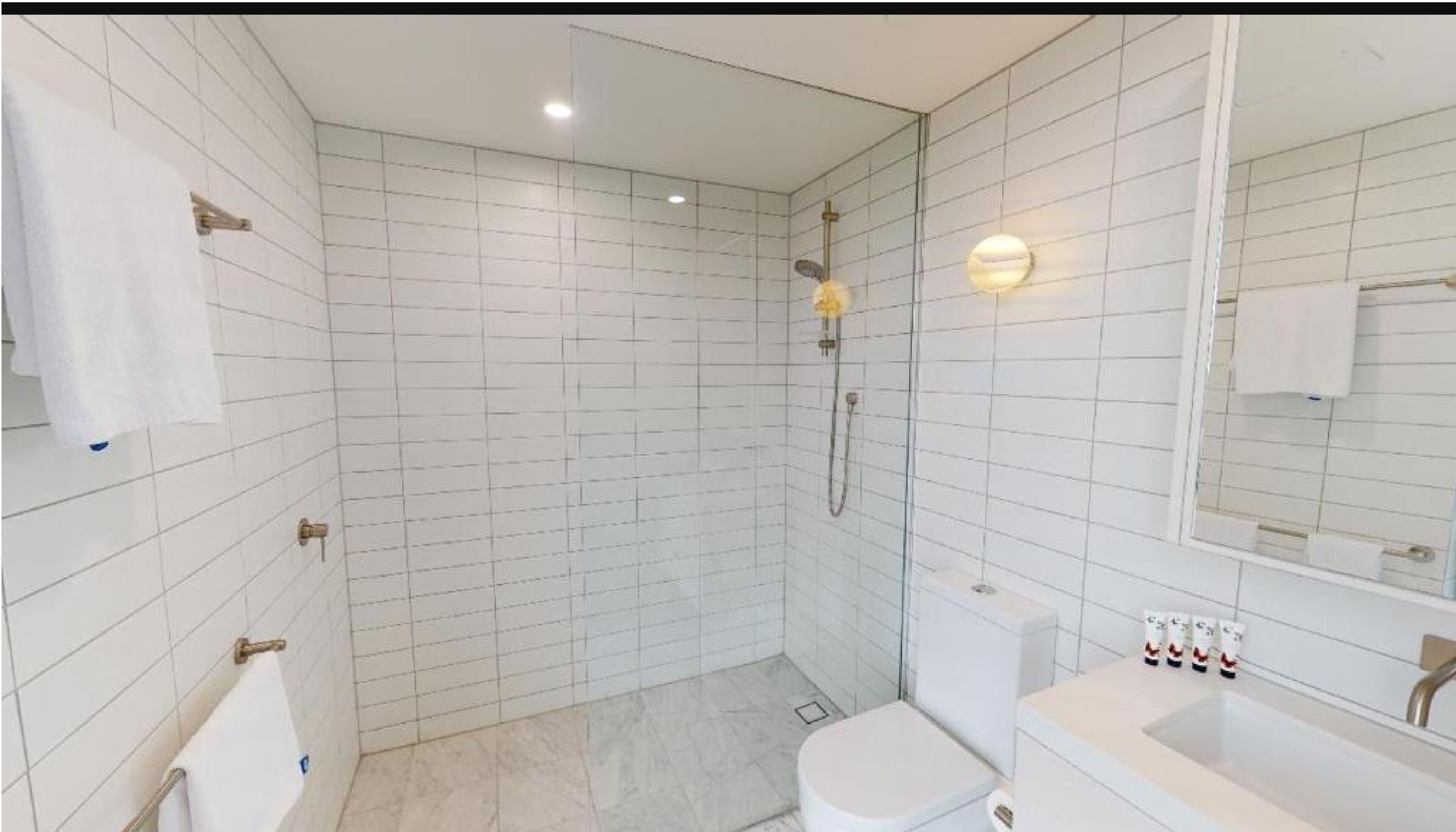 Un baño moderno con paredes de azulejos blancos, cabina de ducha de vidrio, inodoro y lavabo con artículos de aseo.
