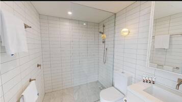 Un baño moderno con paredes de azulejos blancos, cabina de ducha de vidrio, inodoro y lavabo con artículos de aseo.