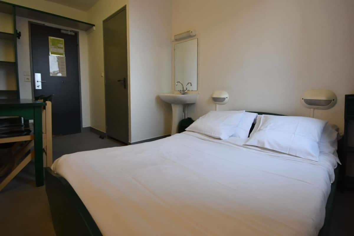 A hotel room with a bed, a desk, a chair, a sink, and a mirror.