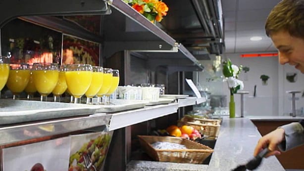 A kitchen area with a counter displaying sliced ham, a fruit basket, and a juice dispenser.