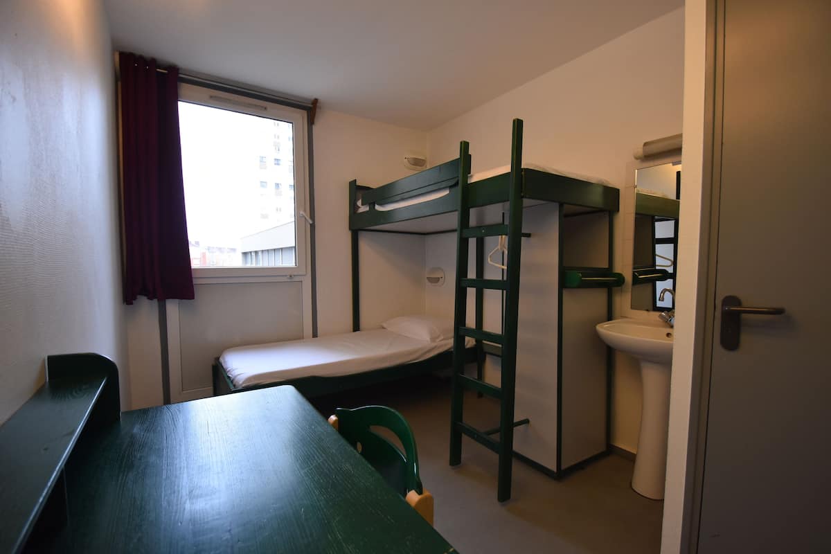 A dormitory room with bunk beds, a desk, a chair, a window with curtains, and a sink.