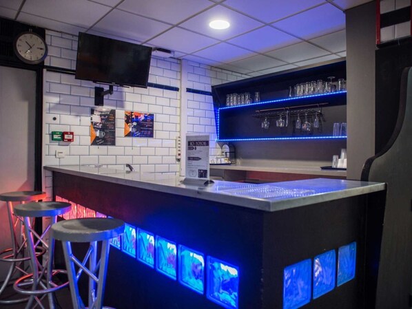 A bar area with a counter, stools, a TV mounted on the wall, and shelves with glasses and bottles.