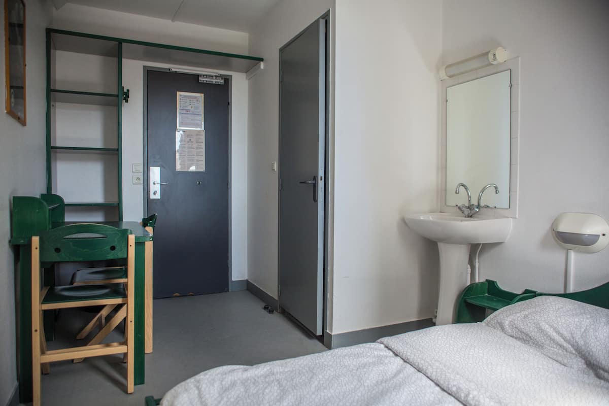 A hotel room with a bed, a green chair, a desk, a wardrobe, and a bathroom with a sink and mirror.