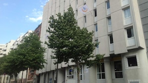 A multi-story building with a modern architectural style, featuring a series of windows and balconies. There are flags hanging from the building, and a tree is visible in front of it.