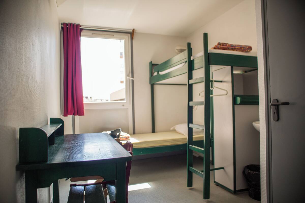 A bunk bed room with a desk and chair, a window with a curtain, and a sink.