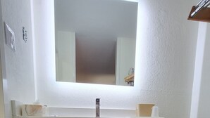 Une salle de bain avec un meuble-lavabo en bois foncé, un lavabo blanc et un miroir au-dessus. Des serviettes sont rangées sous le meuble-lavabo, et une poubelle se trouve sur le côté.