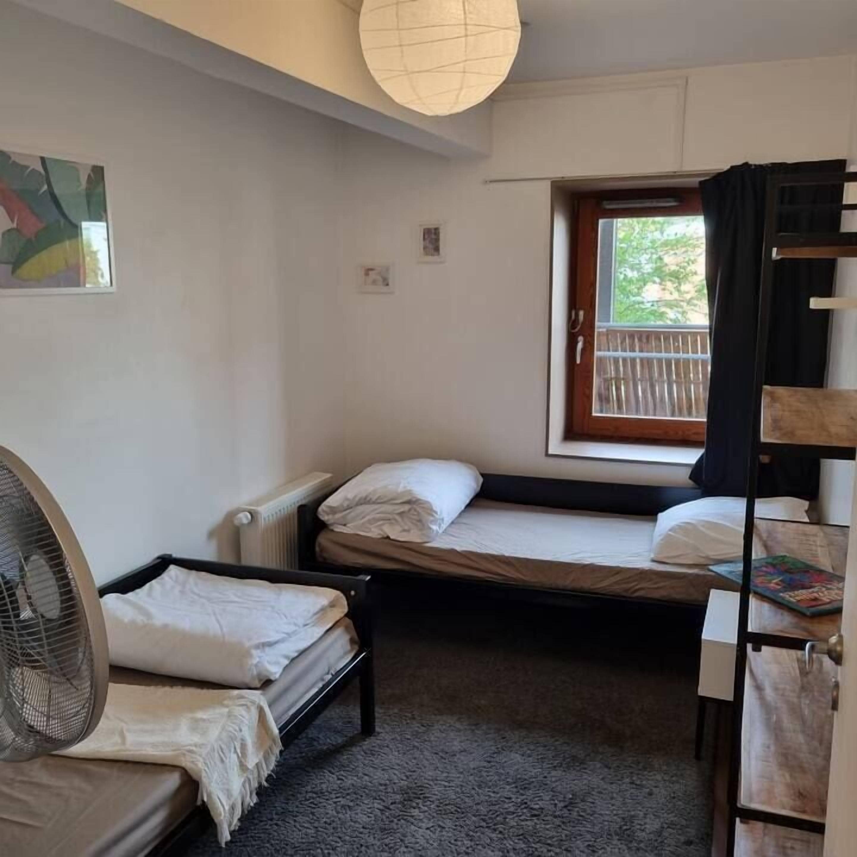 Een kleine, compacte kamer met twee stapelbedden, een enkel bed, een radiator, een ventilator en een raam met uitzicht op buiten.