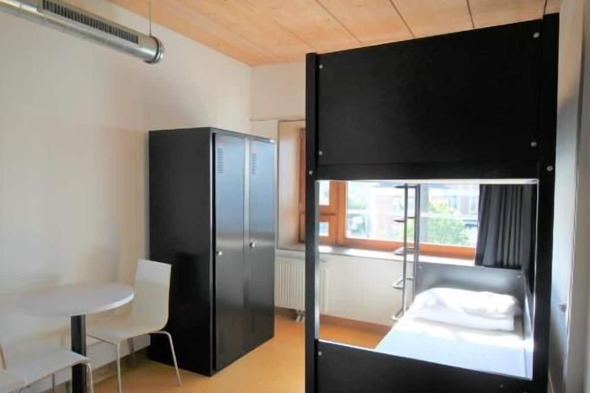 Una habitación compacta con una cama litera, una mesita pequeña, un refrigerador y una ventana con vistas.