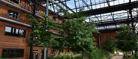 Edifício moderno com teto de vidro e fachada de madeira, apresentando uma passarela para pedestres e vegetação.
