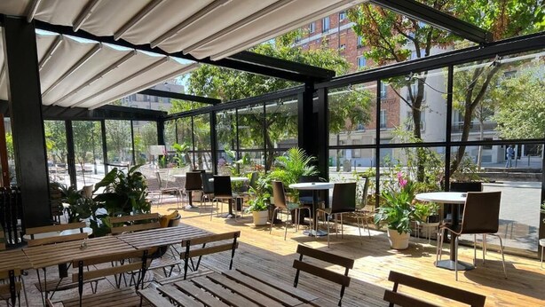 Un café al aire libre con mesas y sillas de madera, grandes ventanales y plantas en macetas.