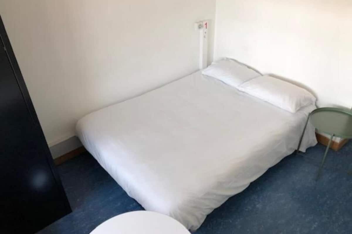 Una habitación pequeña con una cama individual, alfombra azul, ropa de cama blanca y una mesita de noche verde.
